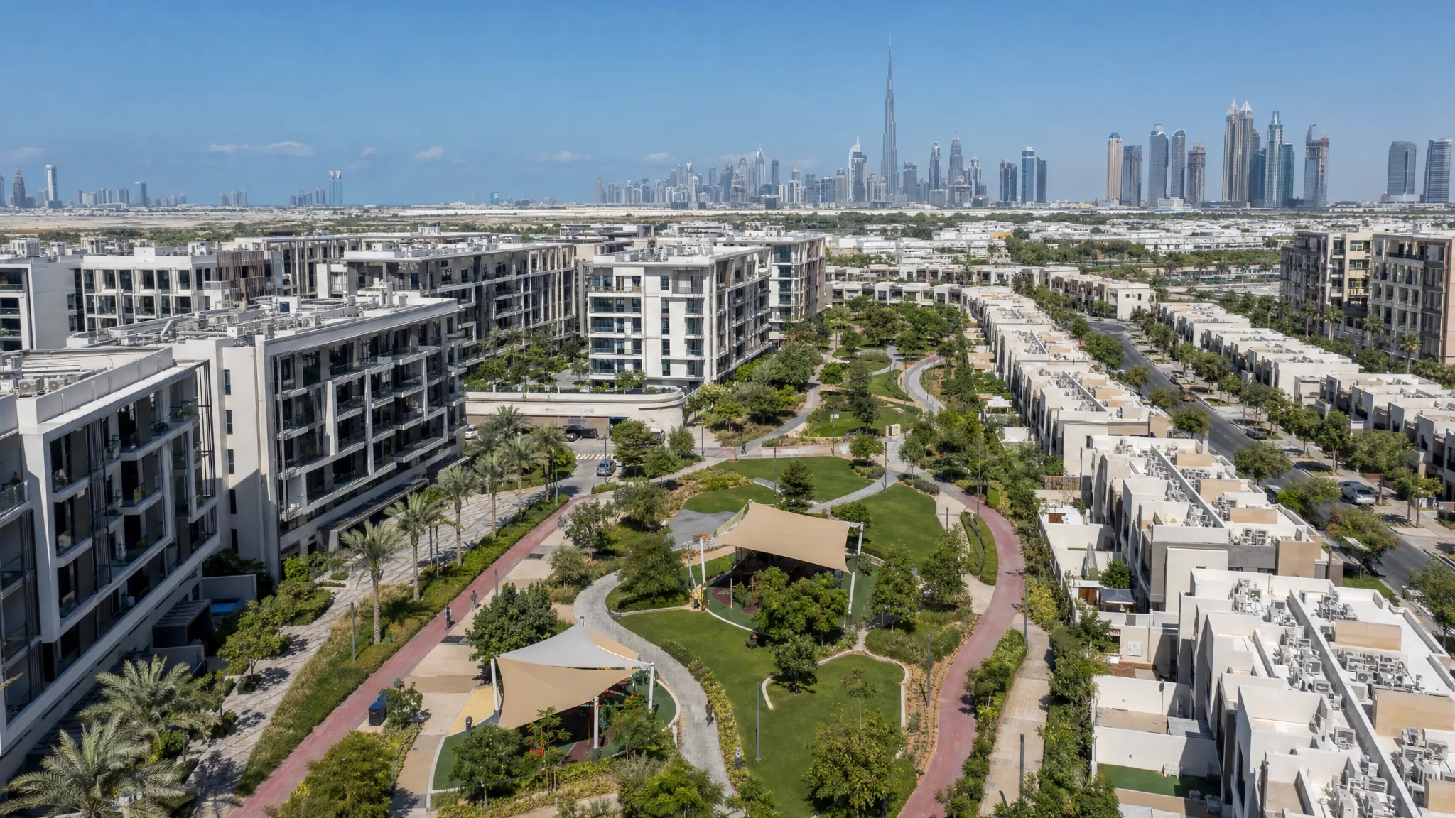 A modern Dubai residential community with mid-rise apartment buildings, townhouses, landscaped walking paths, shaded parks, and the distant city skyline under a clear sky, viewed from an overhead angle with the community layout spread across the frame.